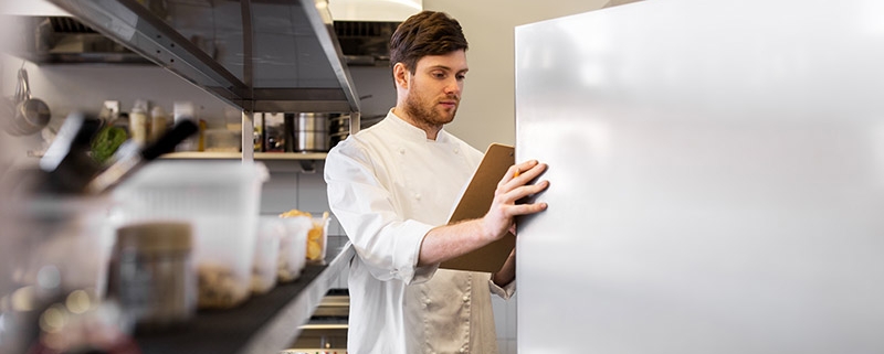 A chef consulting a clipboard while looking through a refrigerator